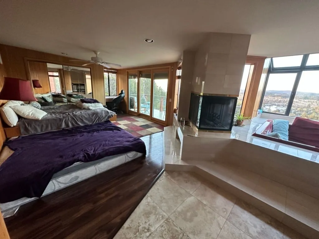 A spacious bedroom with a fireplace, mirrored wall, and windows onto the hills