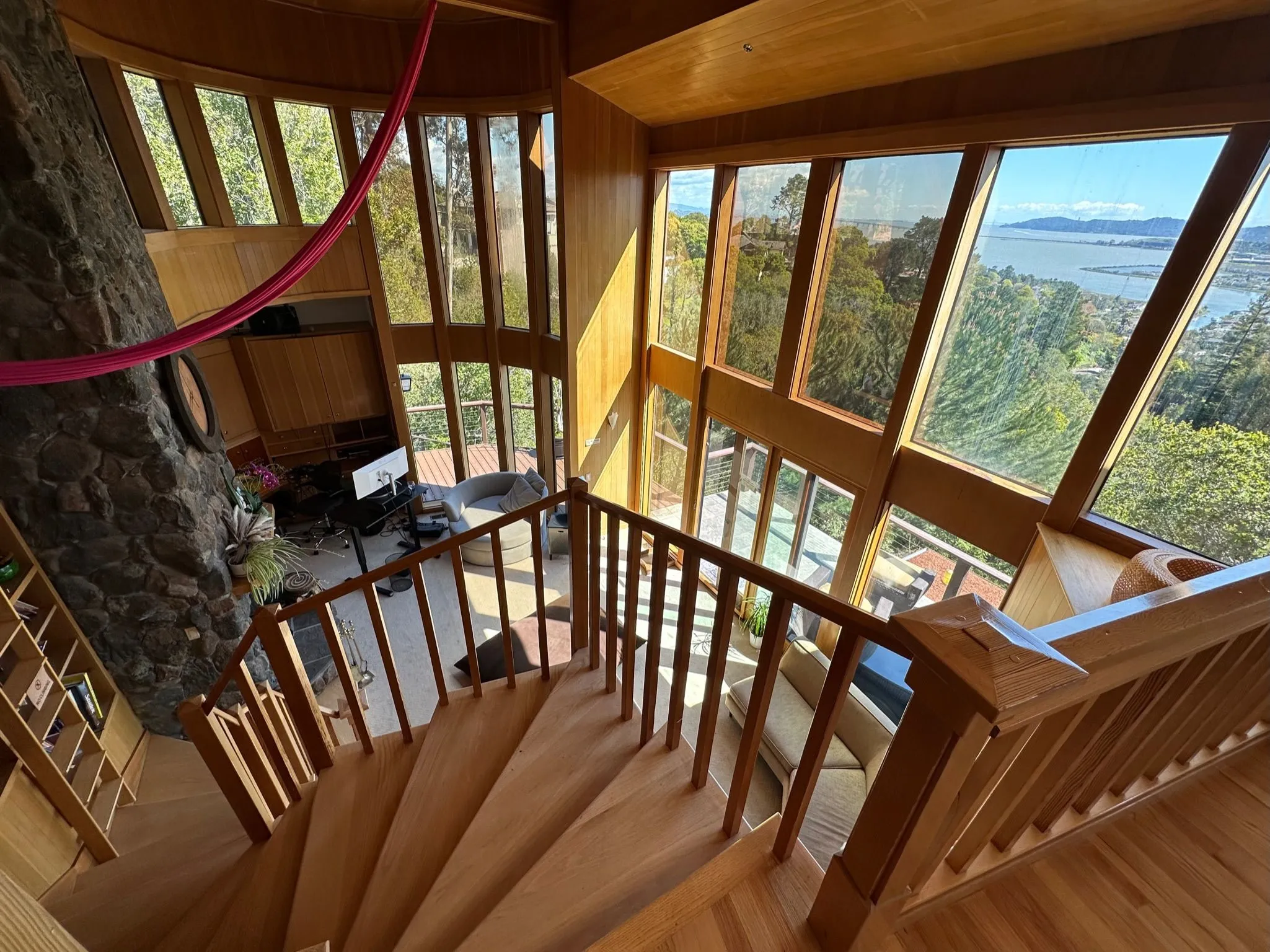 A curved wooden staircase descending past tall windows with a view of the bay